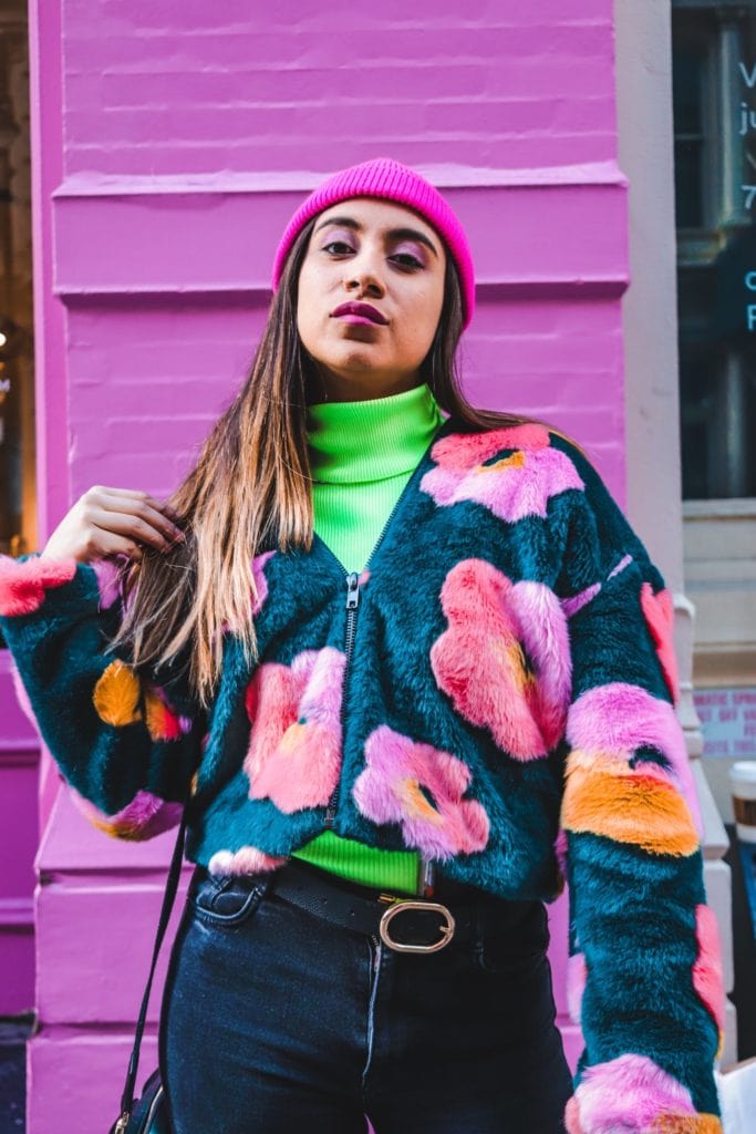 Junge Frau mit auffälligem Style posiert vor einer pinken Wand. Sie trägt einen neongrünen Rollkragen-Pullover, darüber eine halb geöffnete dunkle Jacke mit großen pinken Blumen, dazu einen Beanie. Vintage ist ein Modetrend in ganz Deutschland, besonders aber in Berlin.