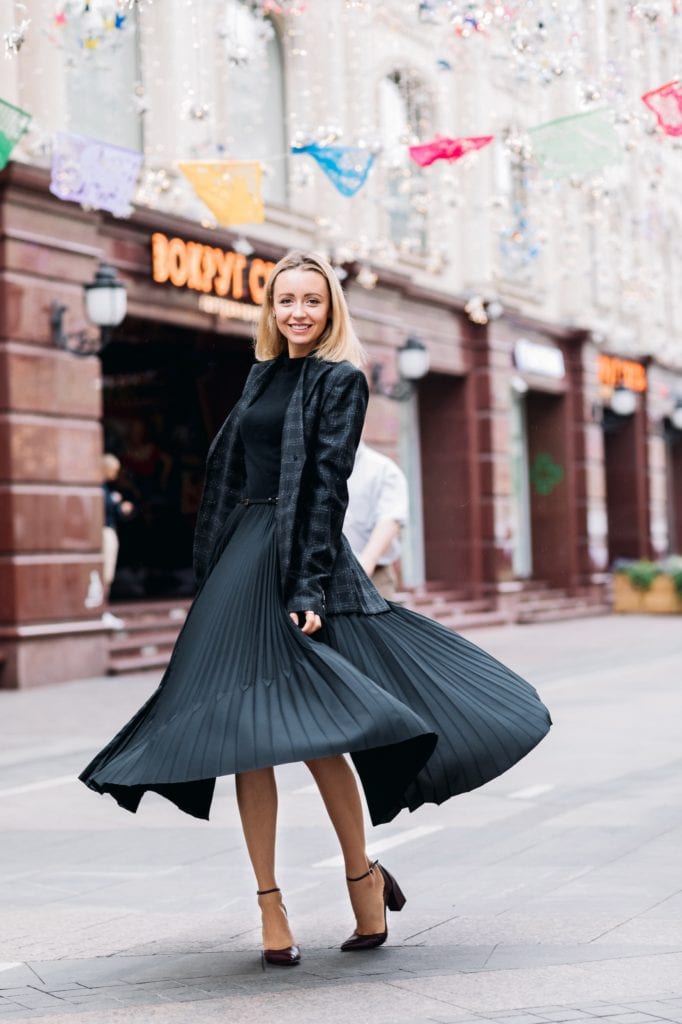 Eine blonde Frau trägt hochhackige schwarze Schuhe, ein schwarzes klassisch-elegantes Kleid und darüber einen schwarzen Blazer. Sie steht in einer Fußgängerzone und lächelt, das Kleid umhüllt ihre Beine wie ein Zelt.