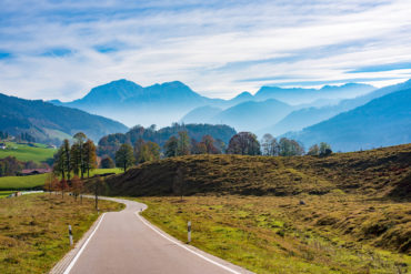 Kurvige Strasse im Bergtal im Herbst. Landschaftliche Ansicht der Fahrbahn, sanft abfallende Hügel, Wiesen mit gelbem Gras, goldenem Sonnenlicht und malerischen Bergen im Hintergrund laden zu einem Roadtrip durch Deutschland ein