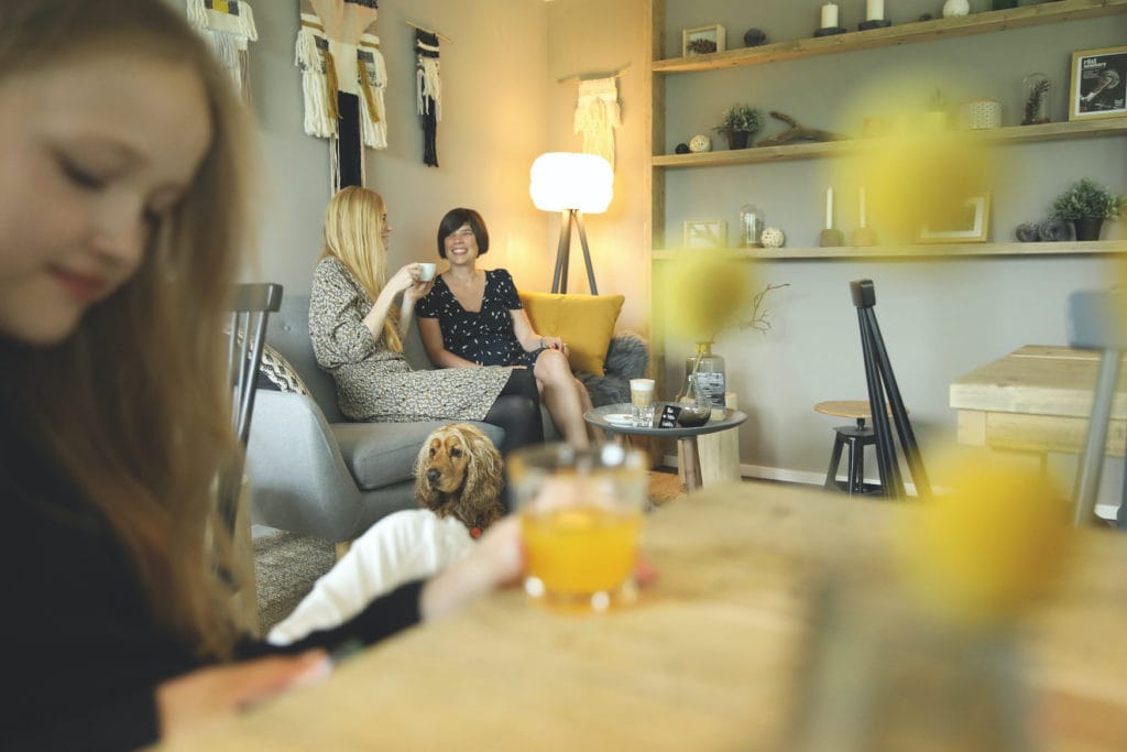 Zwei Frauen sitzen auf einer Couch im Cafe und plauschen, während ein anderer weiblicher Gast vorne liest 