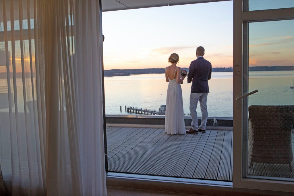 Hochzeitspaar steht auf Balkon in Penthouse Suite in Glücksburg und blickt auf Sonnenuntergang