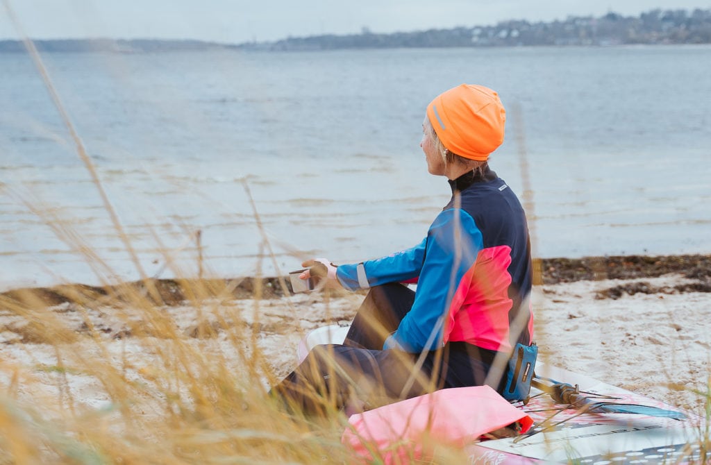 Frau sitzt an Ostsee-Strand und trinkt einen warmen Tee bevor sie zum Stand Up Paddling aufs Wasser geht