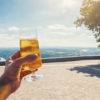 Junger Mann hält Glas mit Bier in die Kamera vor sonnigen Aussichten