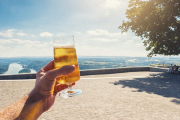 Junger Mann hält Glas mit Bier in die Kamera vor sonnigen Aussichten