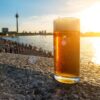 Ein Glas Altbier steht auf einem Mäuerchen oberhalb der Rheinpromenade in Düsseldorf im Sonnenschein. Im Hintergrund sind Rhein und der Düsseldorfer Fernsehturm zu sehen.