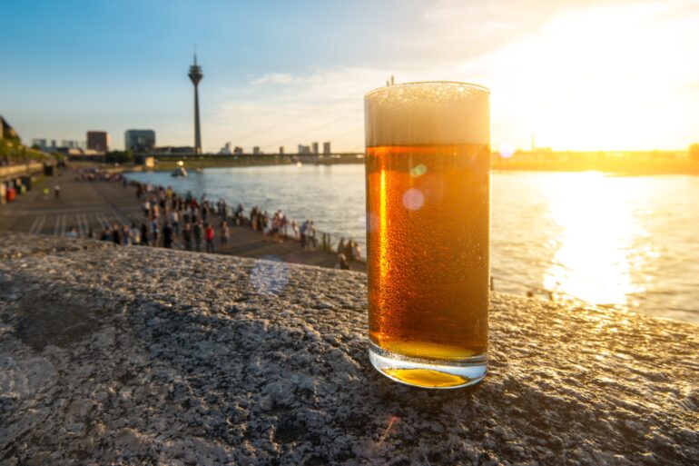 Ein Glas Altbier steht auf einem Mäuerchen oberhalb der Rheinpromenade in Düsseldorf im Sonnenschein. Im Hintergrund sind Rhein und der Düsseldorfer Fernsehturm zu sehen.