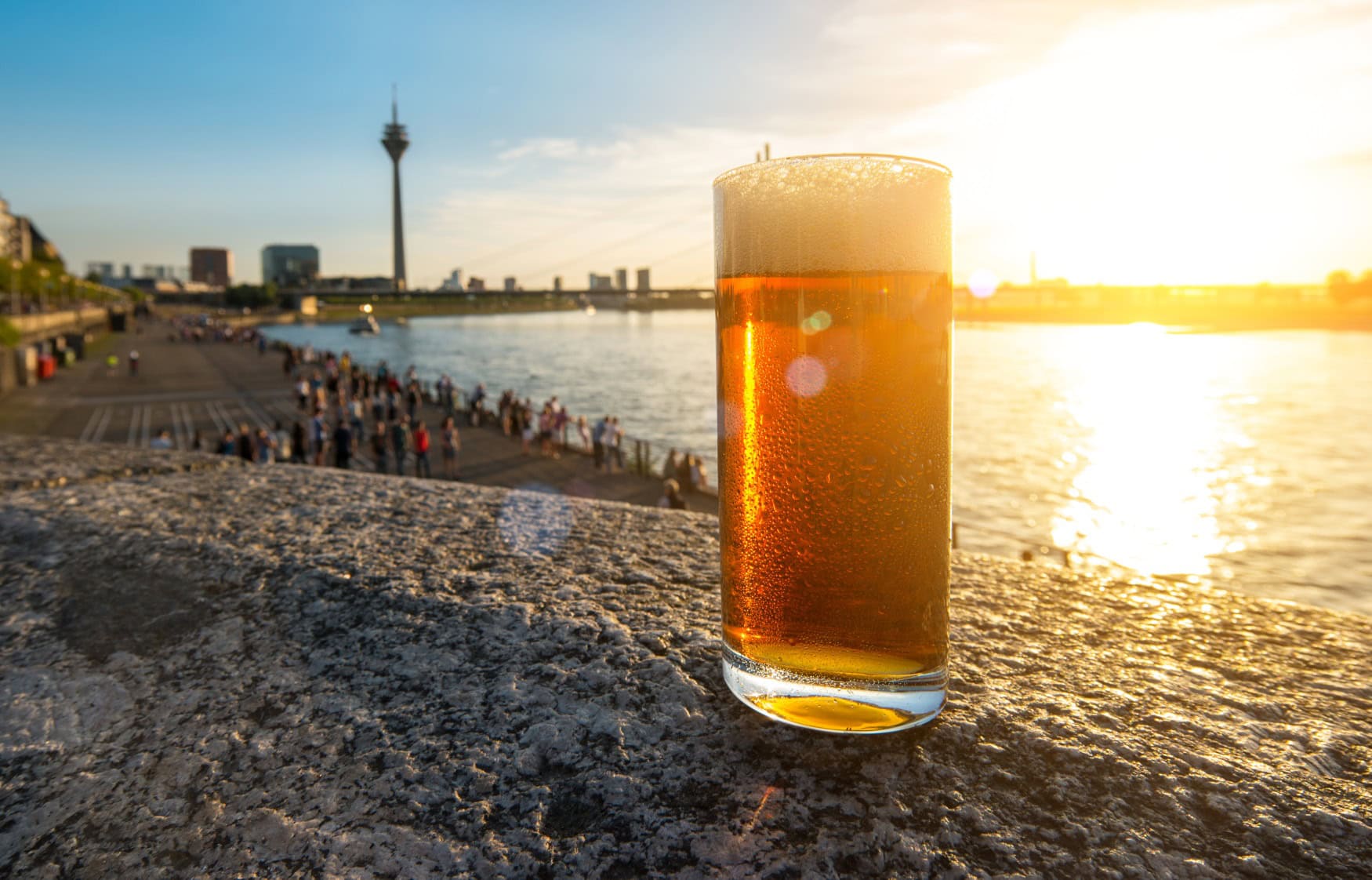 Ein Glas Altbier steht auf einem Mäuerchen oberhalb der Rheinpromenade in Düsseldorf im Sonnenschein. Im Hintergrund sind Rhein und der Düsseldorfer Fernsehturm zu sehen.