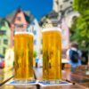 Zwei gefüllte Gläser Kölsch auf einem Holztisch im Freien. Im Hintergrund sind unscharf die bunten Häuser am Alter Markt in Köln zu erkennen