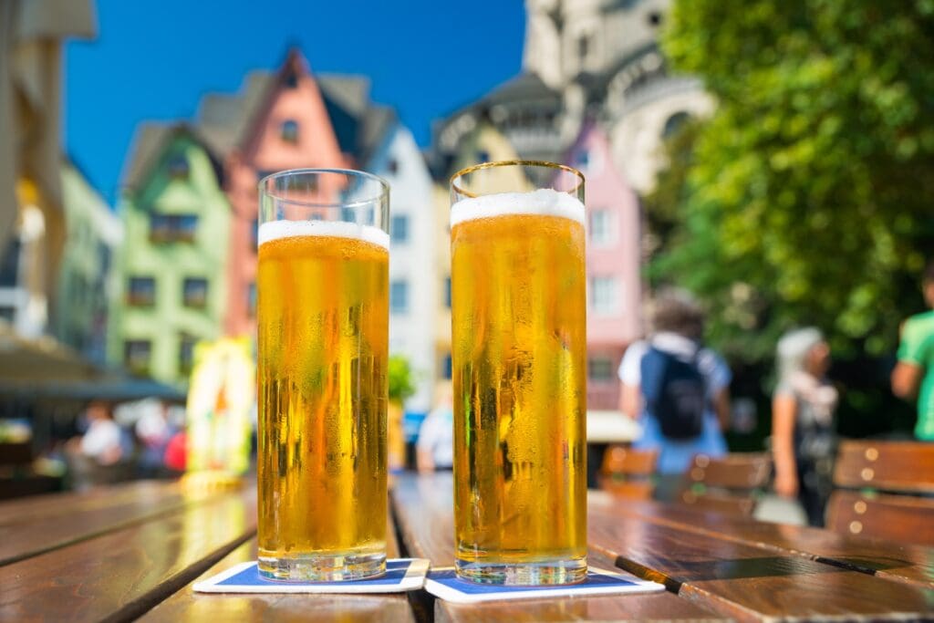 Zwei gefüllte Gläser Kölsch auf einem Holztisch im Freien. Im Hintergrund sind unscharf die bunten Häuser am Alter Markt in Köln zu erkennen