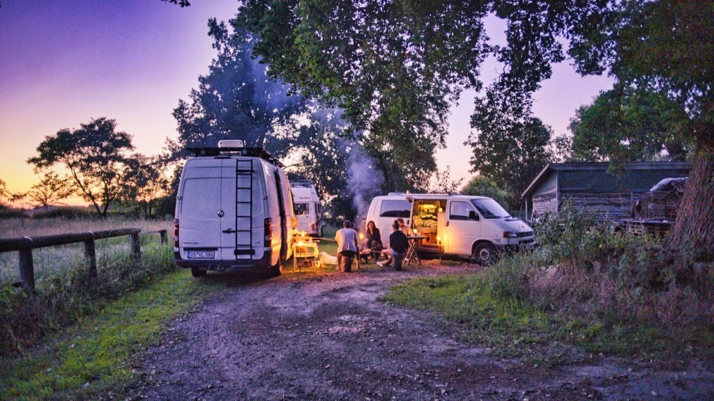 Menschen machen ein Lagerfeuer vor ihren Wohnmobilen auf einem Bauernhof in Deutschland, gebucht haben sie den Stellplatz über Stadt Land Bus Camping