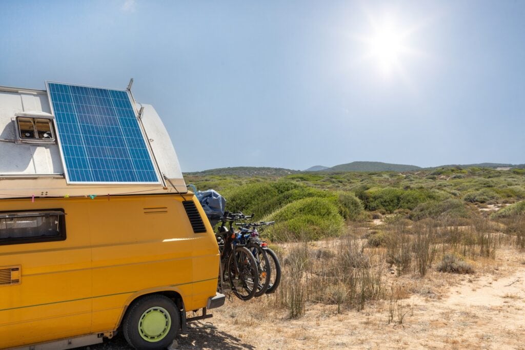 Ein gelber Camping-Bulli mit Solar-Panel auf dem Dach steht in einer sonnigen Dünenlandschaft