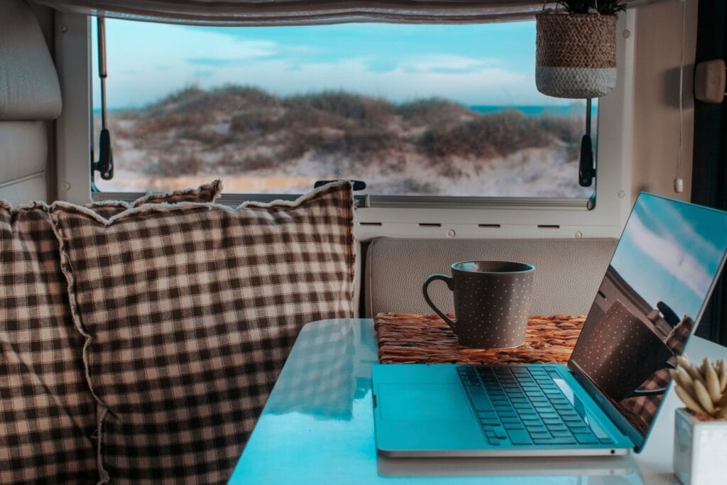 In einem Wohnwagen steht ein Laptop auf dem Tisch, aus dem Fenster heraus sieht man begrünte Dünen