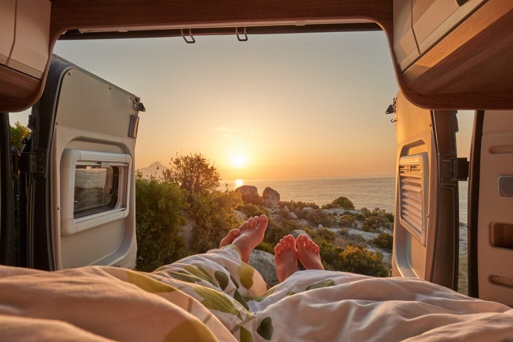 Aus einer geöffneten Hecktür eines Campervans sieht man den Sonnenaufgang über dem Meer. Zwei Menschen strecken ihre Füße aus der Tür