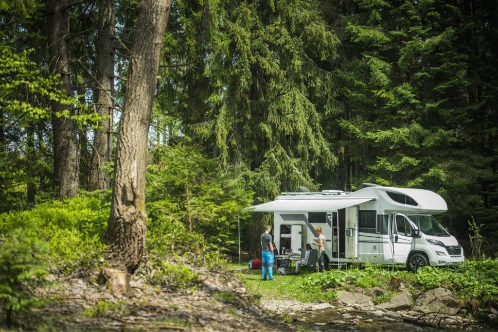 Ein Wohnmobil steht in einem Wald, vier Personen stehen und sitzen unter dem Vordach