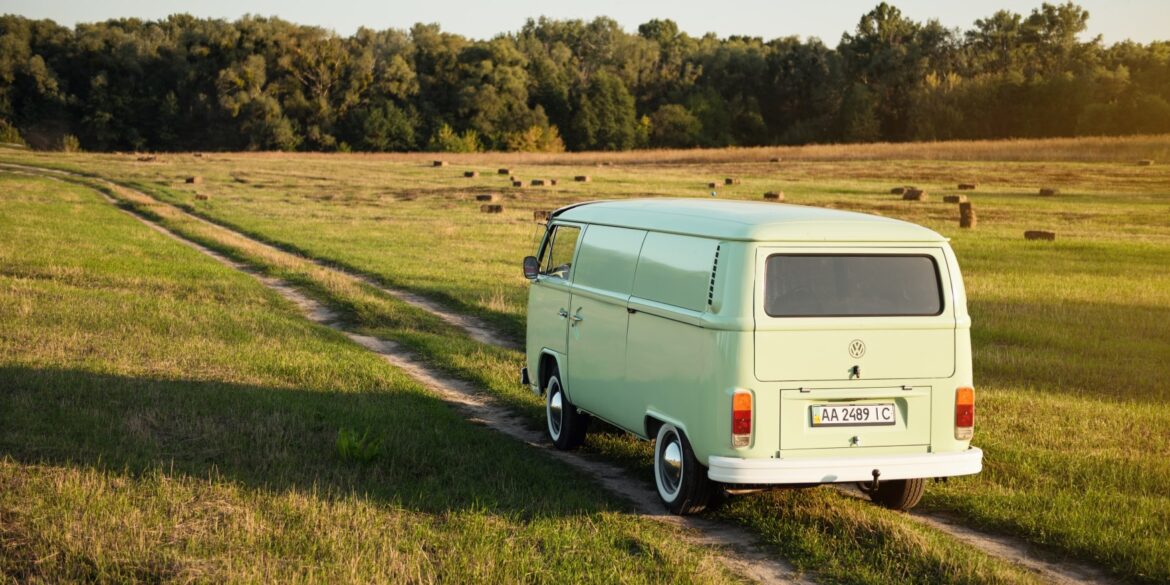 Ein grüner VW Bulli fährt in der Abendsonne über einen Feldweg in Richtung Wald