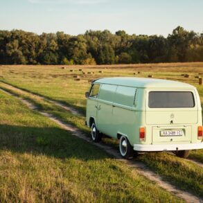 Ein grüner VW Bulli fährt in der Abendsonne über einen Feldweg in Richtung Wald