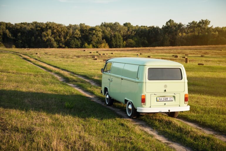 Ein grüner VW Bulli fährt in der Abendsonne über einen Feldweg in Richtung Wald