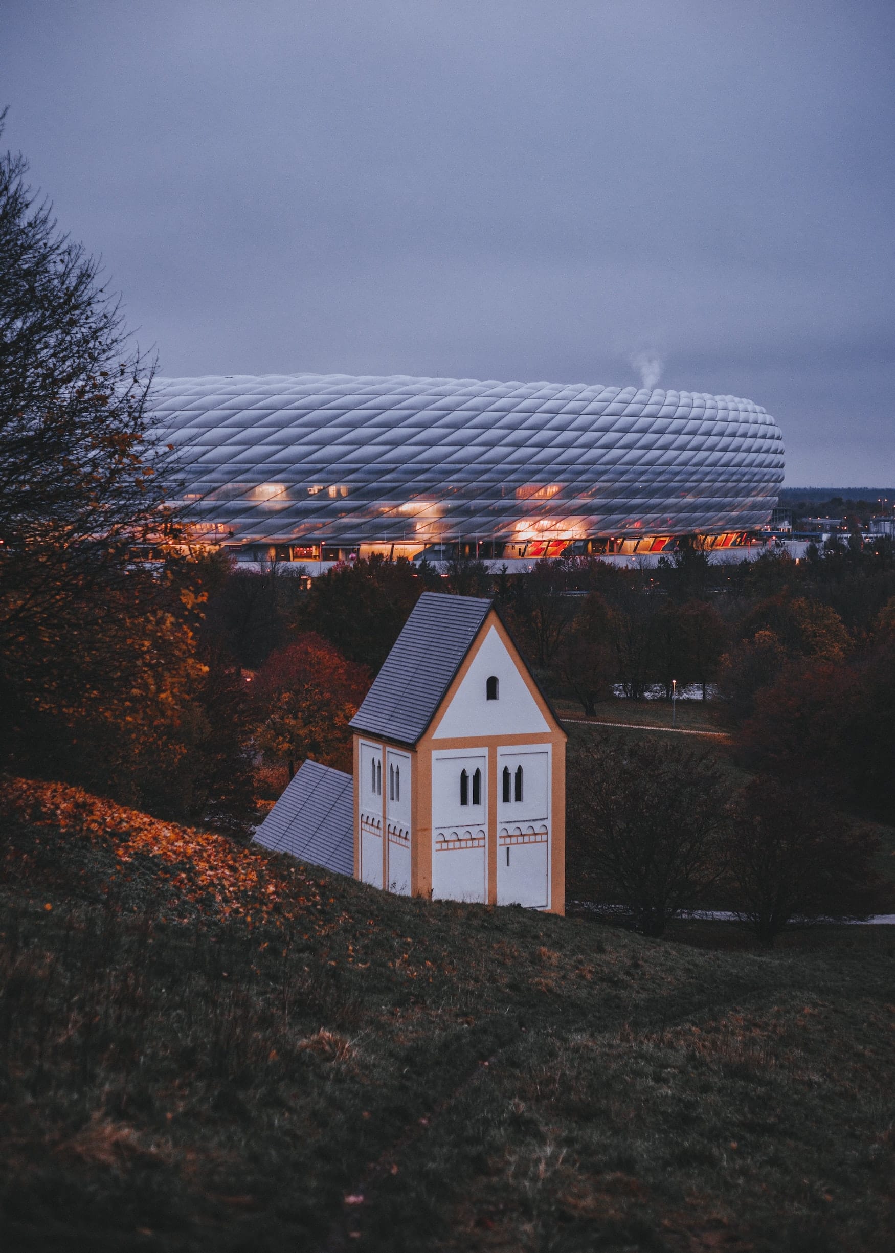 Allianz Arena hinter weiß-gelben Turm in bayerischer Landschaft am Abend