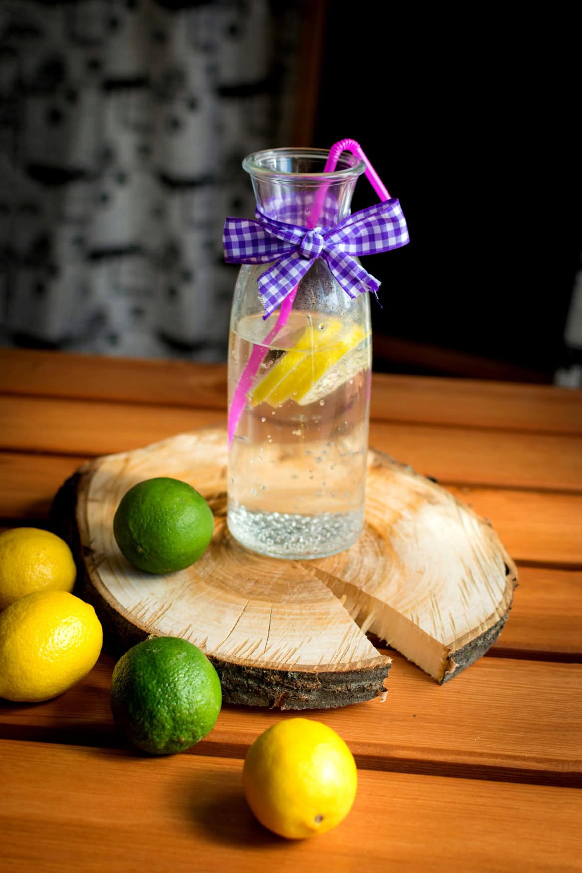 Eine Wasserkaraffe, darin sprudelndes Wasser mit einigen Zitronenscheiben und einem pinken Strohhalm. Um die Flasche ist eine lila-weiß-karierte Schleife gewickelt. Die Flasche steht auf einer Holzscheibe, die wiederum auf einem Holztisch. Um die Flasche herum liegen einige Zitronen und Limetten
