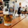 A half full glass of beer with the logo of the Augustiner brewery on the side. It stands on a wooden table, in the background can be seen a beer garden