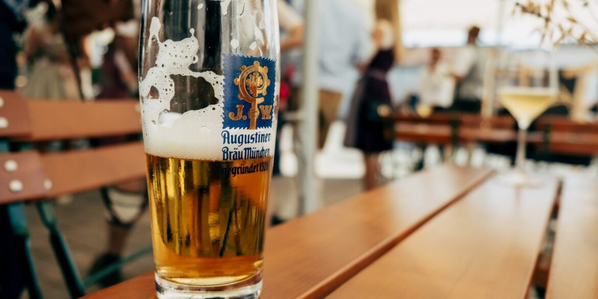 A half full glass of beer with the logo of the Augustiner brewery on the side. It stands on a wooden table, in the background can be seen a beer garden