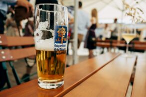 A half full glass of beer with the logo of the Augustiner brewery on the side. It stands on a wooden table, in the background can be seen a beer garden