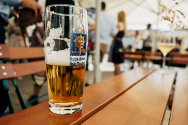 A half full glass of beer with the logo of the Augustiner brewery on the side. It stands on a wooden table, in the background can be seen a beer garden
