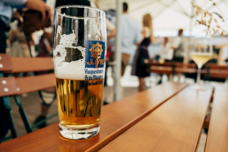 A half full glass of beer with the logo of the Augustiner brewery on the side. It stands on a wooden table, in the background can be seen a beer garden