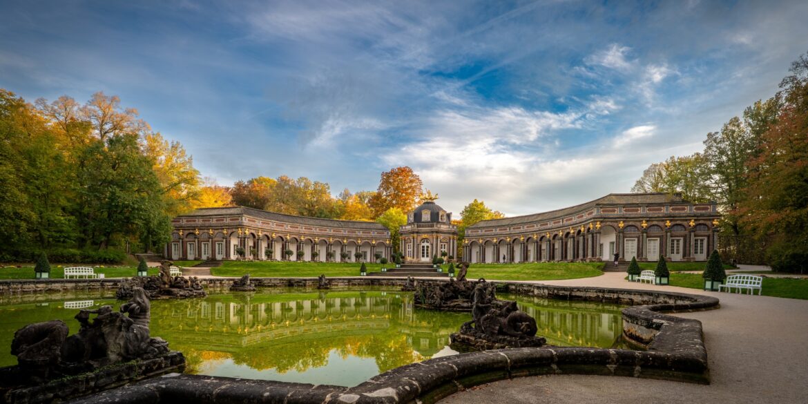 Weitwinkelaufnahme der Eremitage in Bayreuth; im Vordergrund ein breites Wasserbecken, dahinter ein halbrundes Schloss mit mittiger Kuppel.