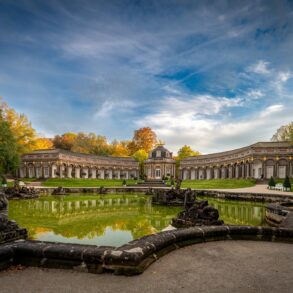Weitwinkelaufnahme der Eremitage in Bayreuth; im Vordergrund ein breites Wasserbecken, dahinter ein halbrundes Schloss mit mittiger Kuppel.