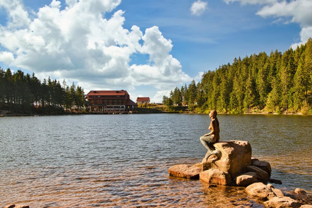 Eine steinerne Nymphenstatue in einem See; im Hintergrund, auf der anderen Seite des Sees, steht ein großes Haus