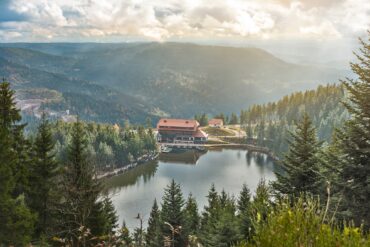 Blick von oben auf einen umwaldeten See mit einem großen Haus am Seeufer. Im Hintergrund sind die Berge des Schwarzwalds zu sehen.
