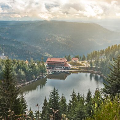 Blick von oben auf einen umwaldeten See mit einem großen Haus am Seeufer. Im Hintergrund sind die Berge des Schwarzwalds zu sehen.