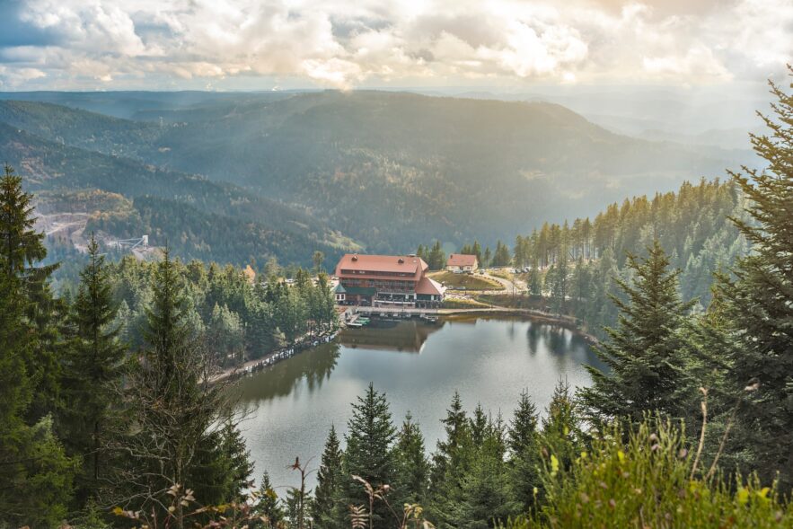Blick von oben auf einen umwaldeten See mit einem großen Haus am Seeufer. Im Hintergrund sind die Berge des Schwarzwalds zu sehen.