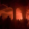 Silhouettes of people dancing in a night club illuminated by red light. In the center of the room are two huge pillars, hinting on a possible industrial past of the building.
