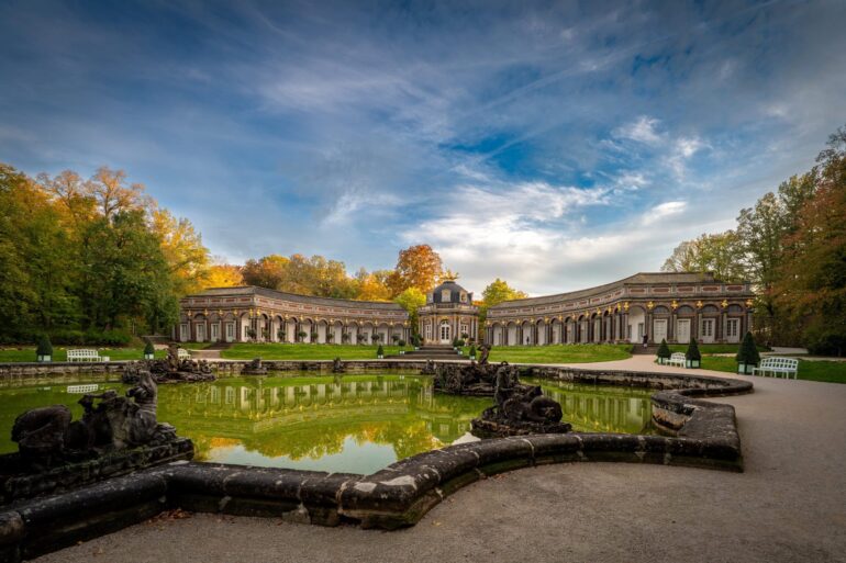 Wide-angle shot of the Hermitage in Bayreuth; in the foreground, a wide water basin; behind it, a semicircular palace with a central dome.