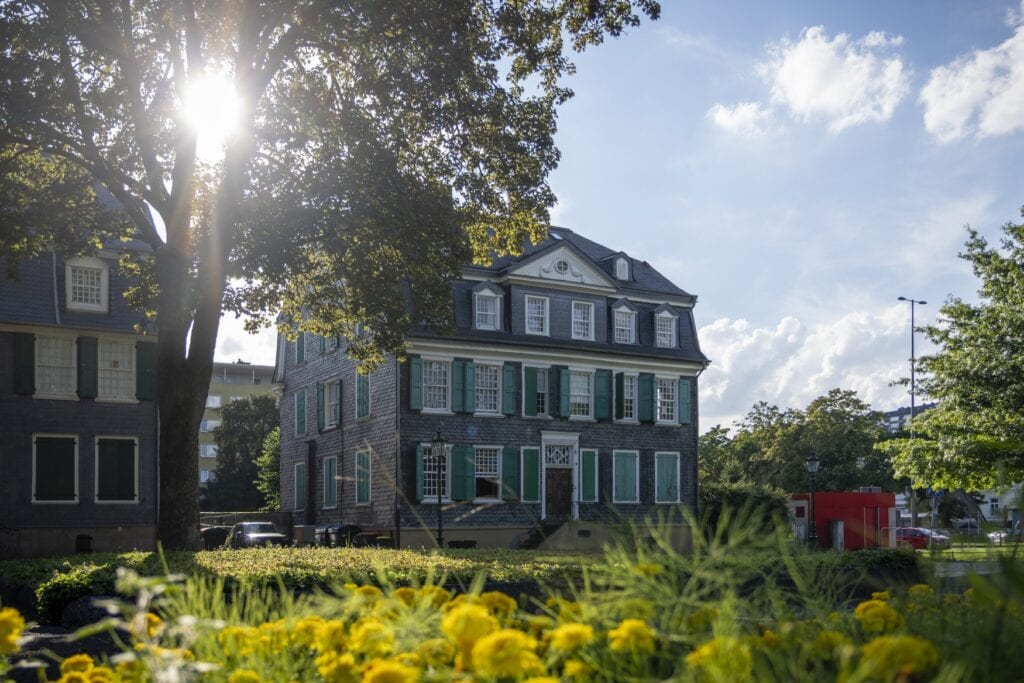 Ein mit schwarzen Schieferplatten verkleidetes Haus mti grünen Fensterläden und weißen Fensterrahmen wird von der Sonne beschienen. Im Vordergrund steht ein hoher Baum, davor eine Wiese mit gelben Blumen