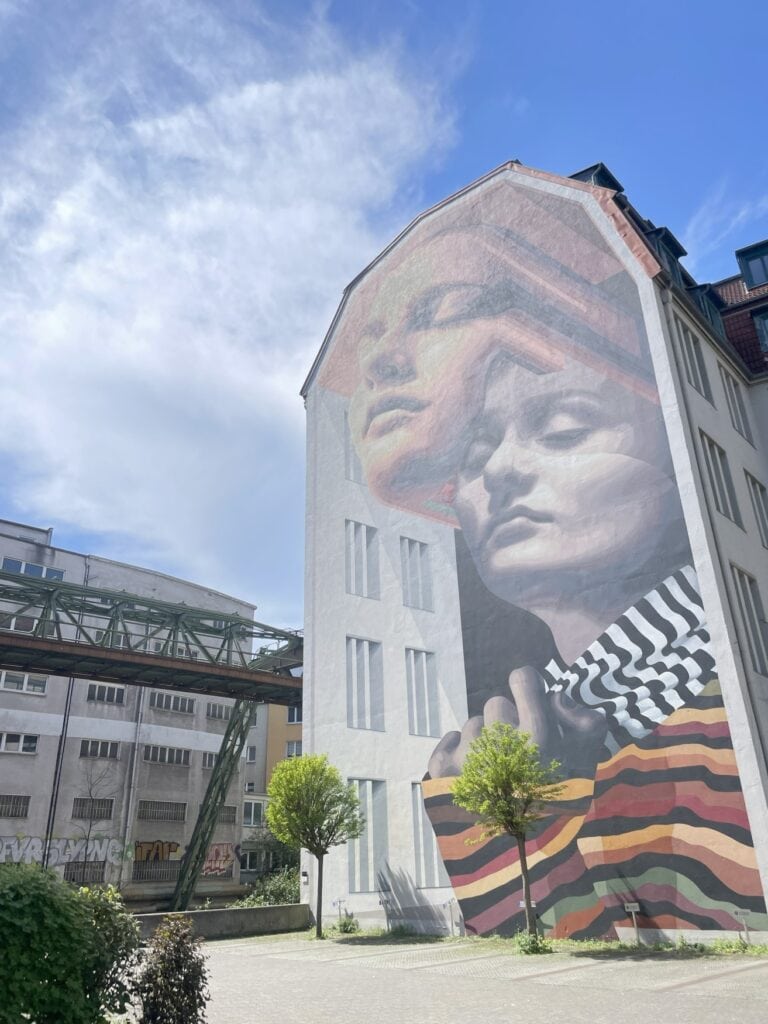 Ein haushohes Wandmural zeigt zwei Frauengesichter und eine gemalte Häuserwand