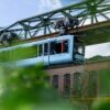 Die Wuppertaler Schwebebahn in hellem Blau fährt von links nach rechts vor einem geziegelten Industriebau vorbei; im Vordergrund etwas Begrünung
