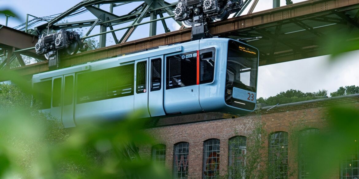 Die Wuppertaler Schwebebahn in hellem Blau fährt von links nach rechts vor einem geziegelten Industriebau vorbei; im Vordergrund etwas Begrünung