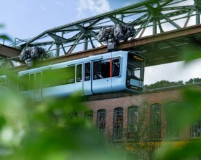 Die Wuppertaler Schwebebahn in hellem Blau fährt von links nach rechts vor einem geziegelten Industriebau vorbei; im Vordergrund etwas Begrünung
