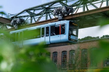 Die Wuppertaler Schwebebahn in hellem Blau fährt von links nach rechts vor einem geziegelten Industriebau vorbei; im Vordergrund etwas Begrünung