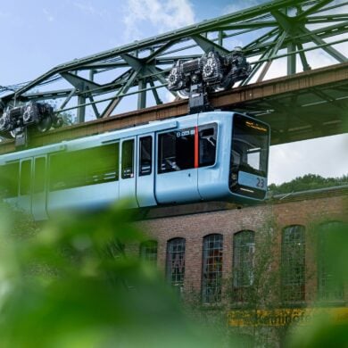 Die Wuppertaler Schwebebahn in hellem Blau fährt von links nach rechts vor einem geziegelten Industriebau vorbei; im Vordergrund etwas Begrünung