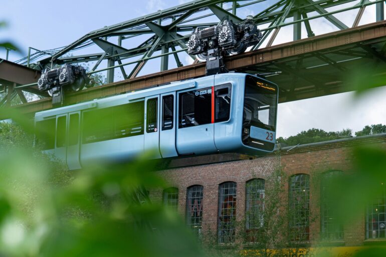 Die Wuppertaler Schwebebahn in hellem Blau fährt von links nach rechts vor einem geziegelten Industriebau vorbei; im Vordergrund etwas Begrünung