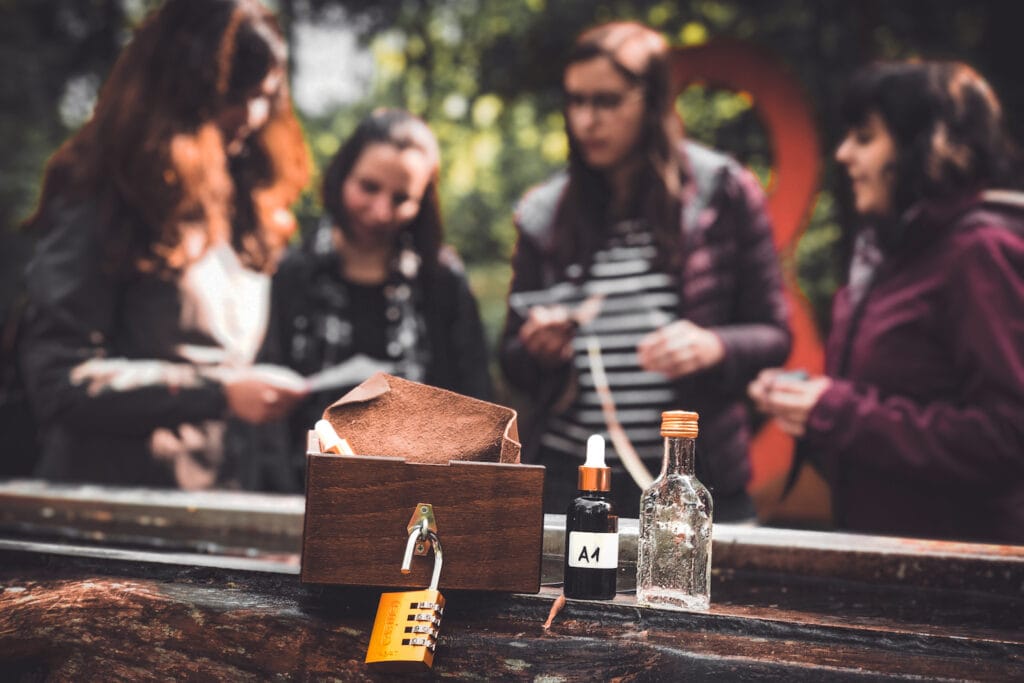 Hinweise eines Rätselspiels: Eine aufgeschlossene Schatulle, ein Medizinfläschchen, eine Wasserflasche; im Hintergrund die Spielerinnen