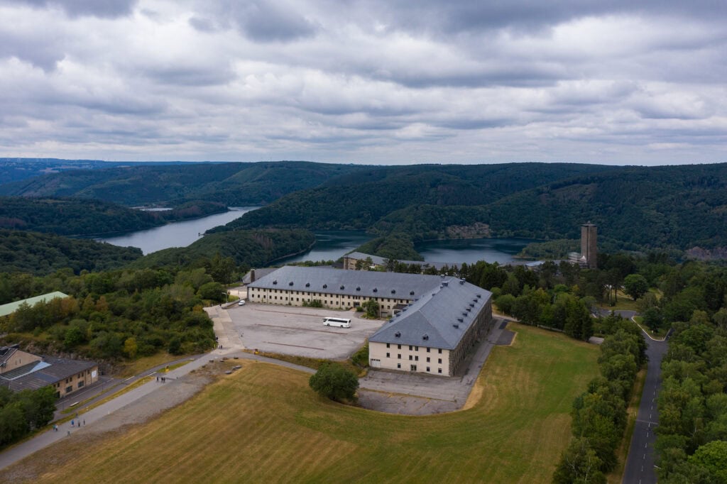 Luftaufnahme der sogenannten "NS-Ordensburg Vogelsang" in der Eifel, ein L-förmiger, kasernenähnlicher Bau auf einer Freifläche in einem Wald. Im Hintergrund hinter dem Gebäude liegt in einem Tal der Rurstausee.