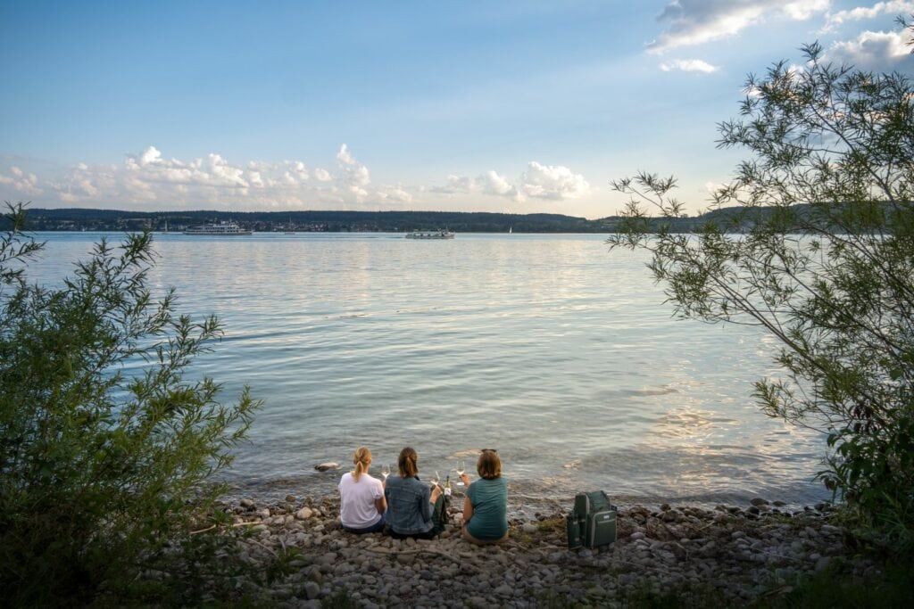 Drei Personen sitzen mit einem Rucksack am Bodensee und trinken Wein
