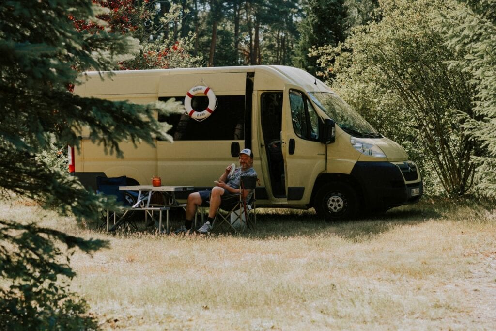 Ein Mann trinkt vor seinem Camper-Van im Wald ein Bier