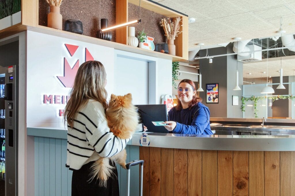 Eine Frau mit Hund auf dem Arm steht mit dem Rücken zum Betrachter an einer Hotelrezeption. Eine Mitarbeiterin reicht ihr über den Tresen ihre Key Card.
