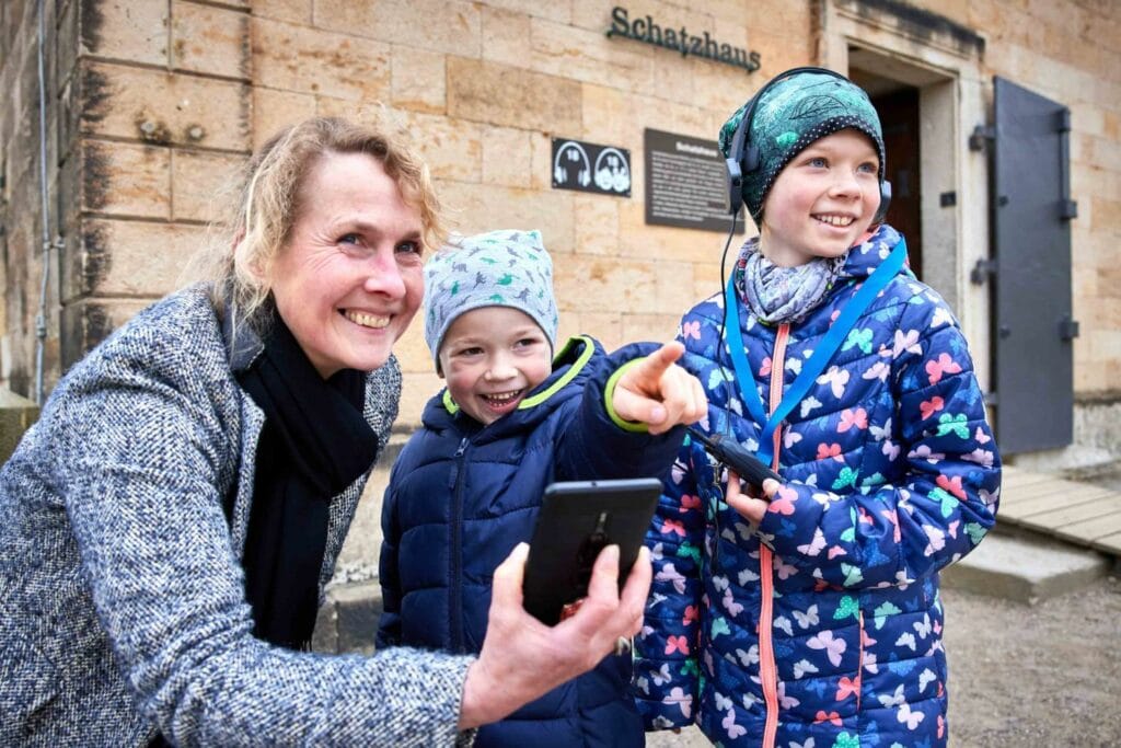 Zwei Kinder und eine erwachsene Frau erkunden die Festung Königstein mit einem Audioguide auf den Ohren